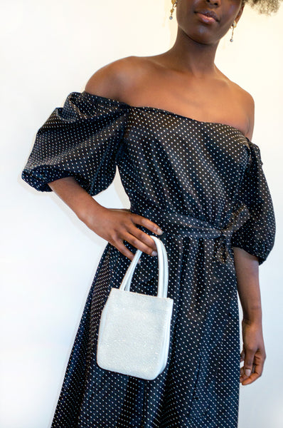 Model wears black puff sleeve dress while holding white leather and glitter handbag